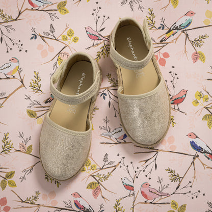 Metallic white linen espadrille with ankle strap and braided jute sole on floral background.