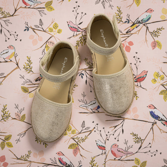 Metallic white linen espadrille with ankle strap and braided jute sole on floral background.
