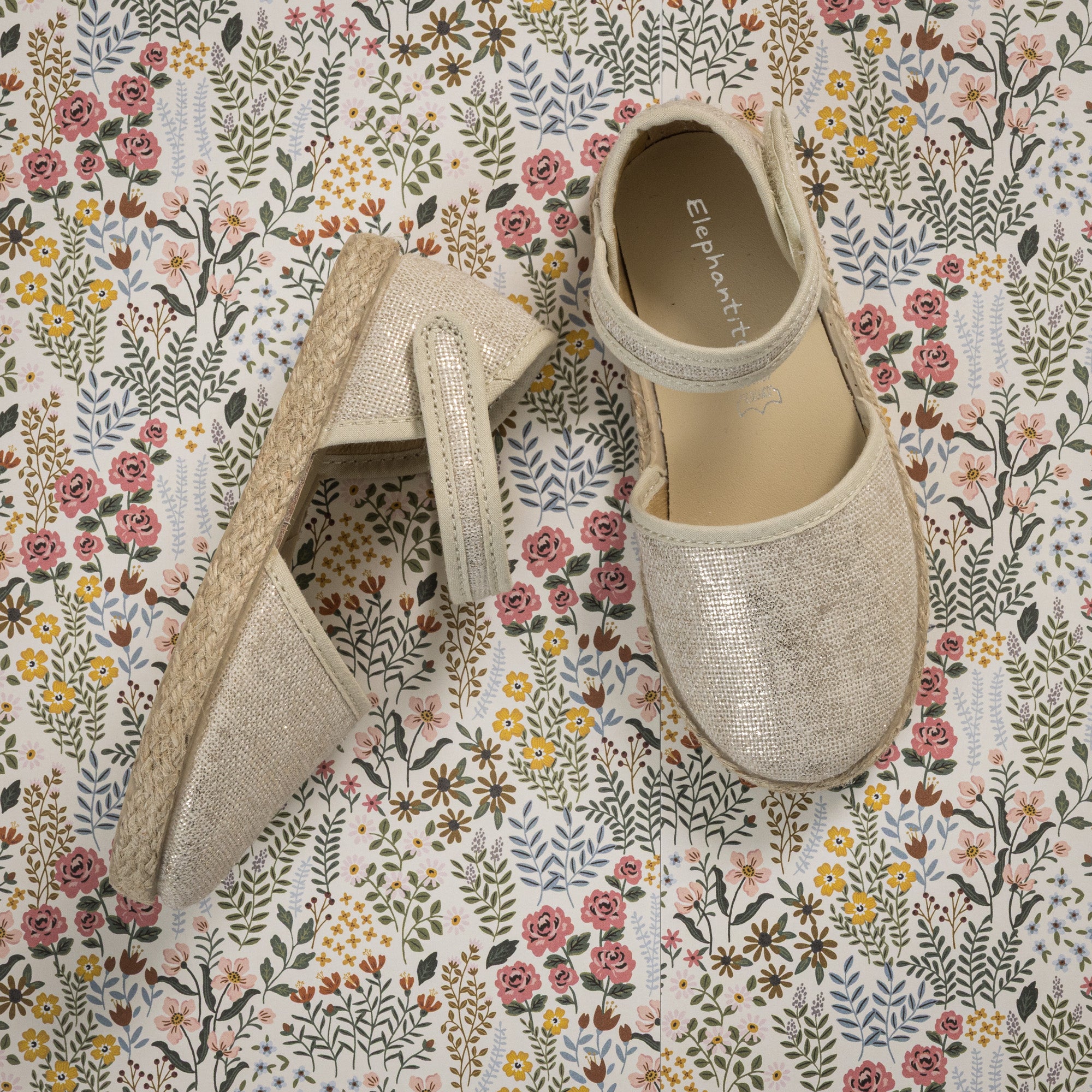Metallic white linen toddler espadrille with ankle strap and jute-wrapped sole on floral background.