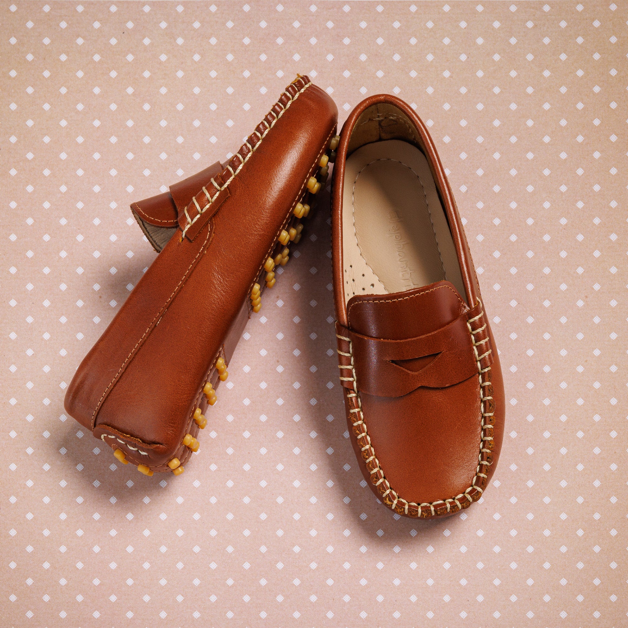 Natural brown leather boys loafers with moccasin stitching and rubber pebble soles on pink polka dot background.