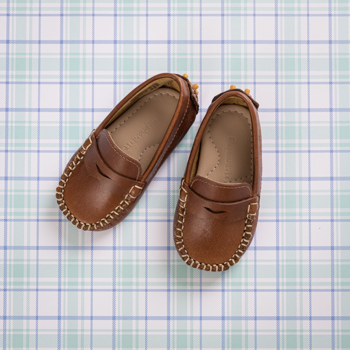 Natural brown toddler moccasin shoes with stitched detailing and penny loafer style on plaid background.