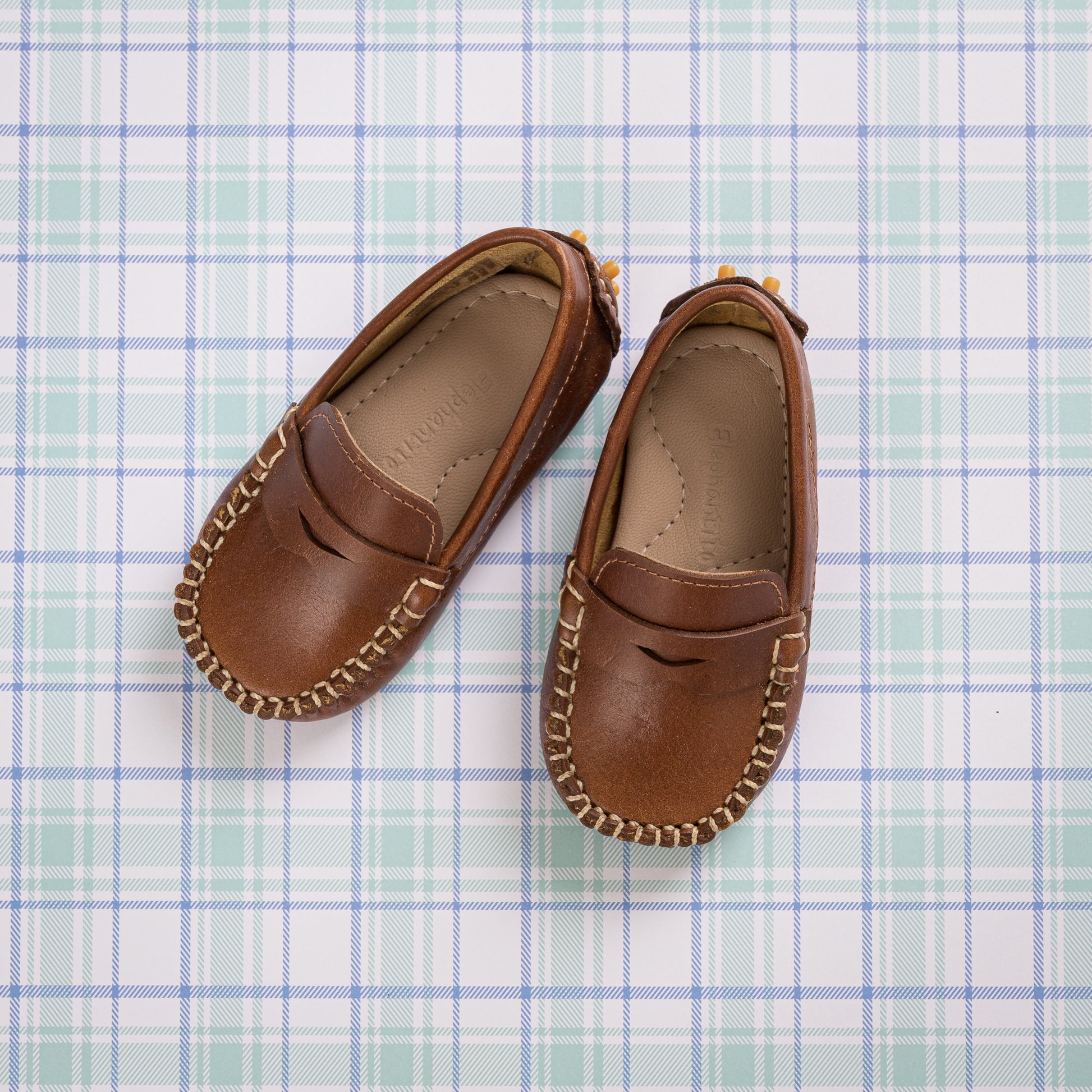 Natural brown toddler moccasin shoes with stitched detailing and penny loafer style on plaid background.