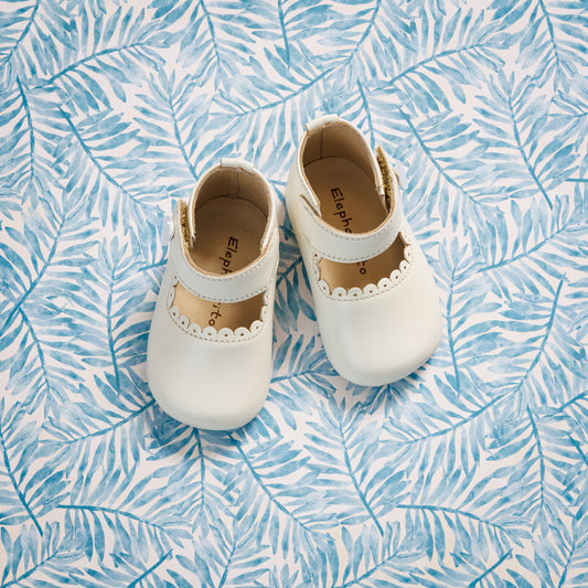 Elephantito - White leather baby Mary Jane shoes with scalloped edges and a buckle strap on blue leaf-patterned fabric background.