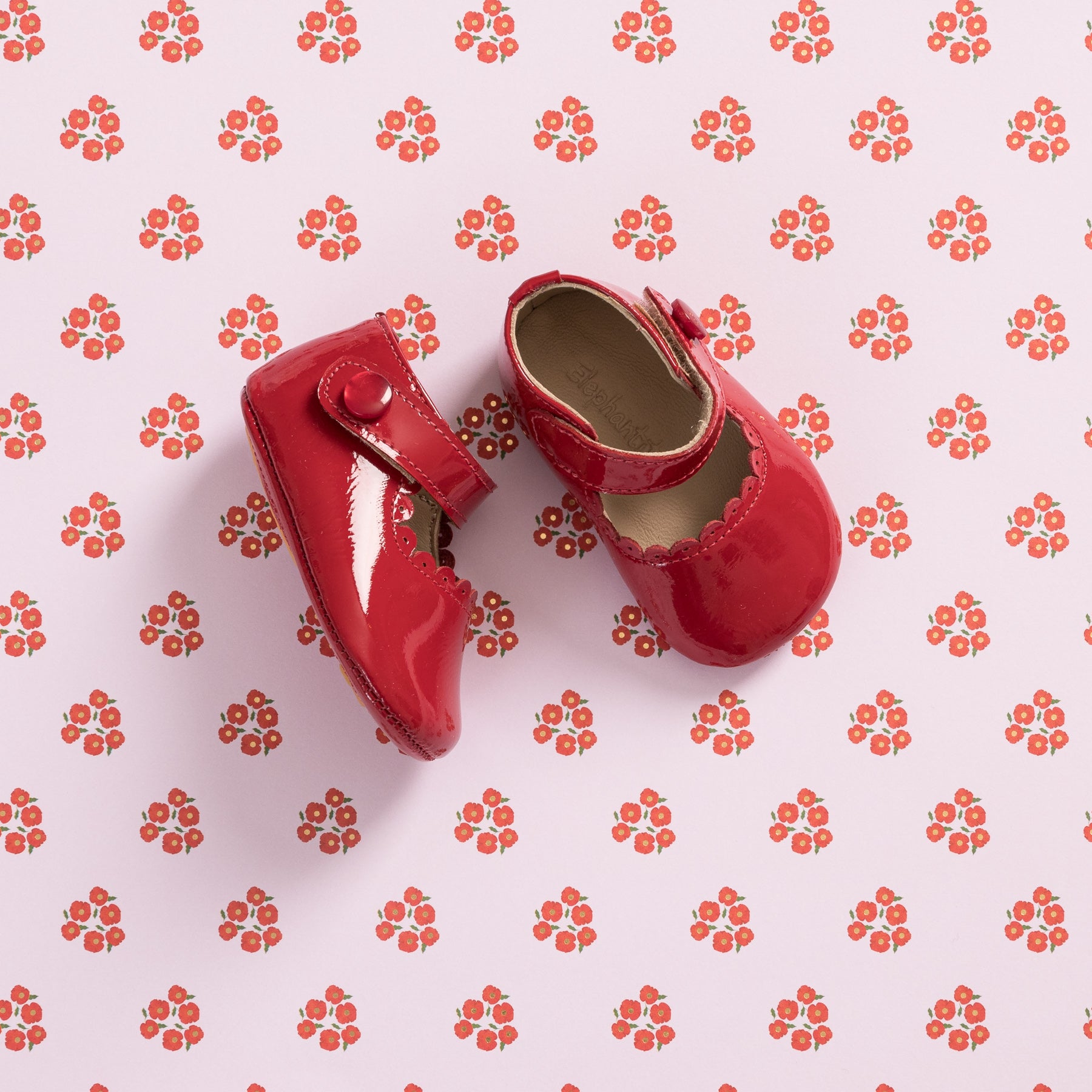 Glossy red patent leather baby Mary Jane shoes with scalloped edges and button strap closure on floral background.