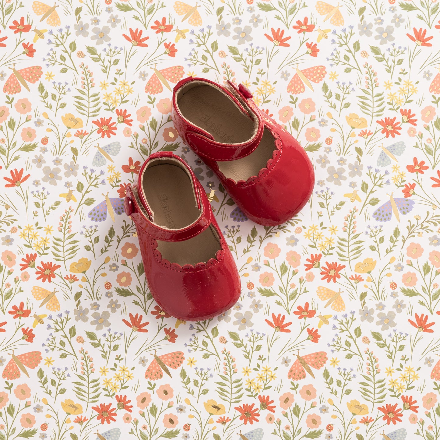 Red patent leather baby Mary Jane shoes with scalloped edges on floral background.
