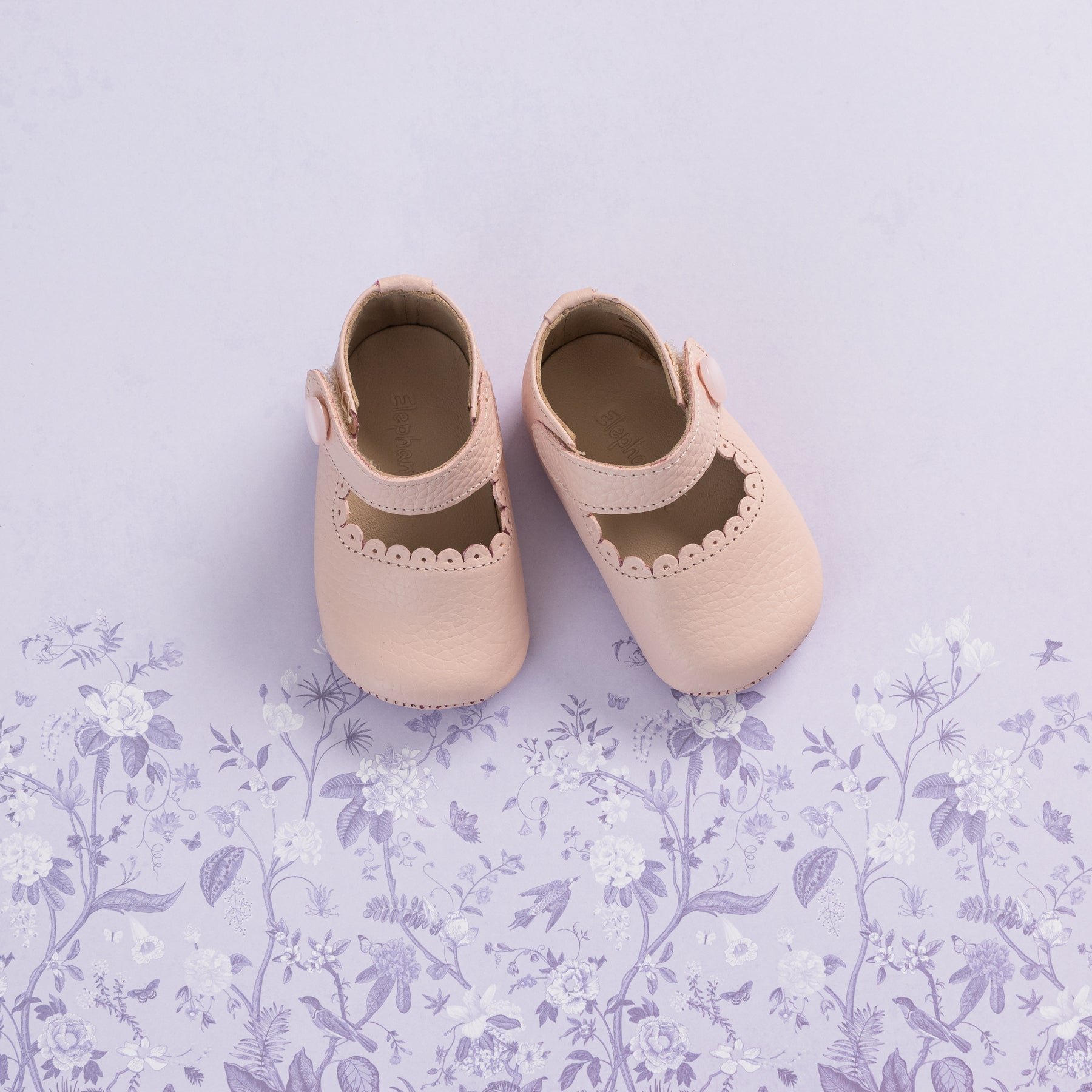 Soft pink baby Mary Jane shoes with scalloped edges and straps on a lavender floral background.