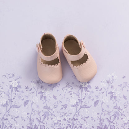 Soft pink baby Mary Jane shoes with scalloped edges and straps on a lavender floral background.