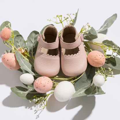 Pink leather baby Mary Jane shoes with scalloped edges and hook-and-loop closures resting on eucalyptus leaves with pastel.