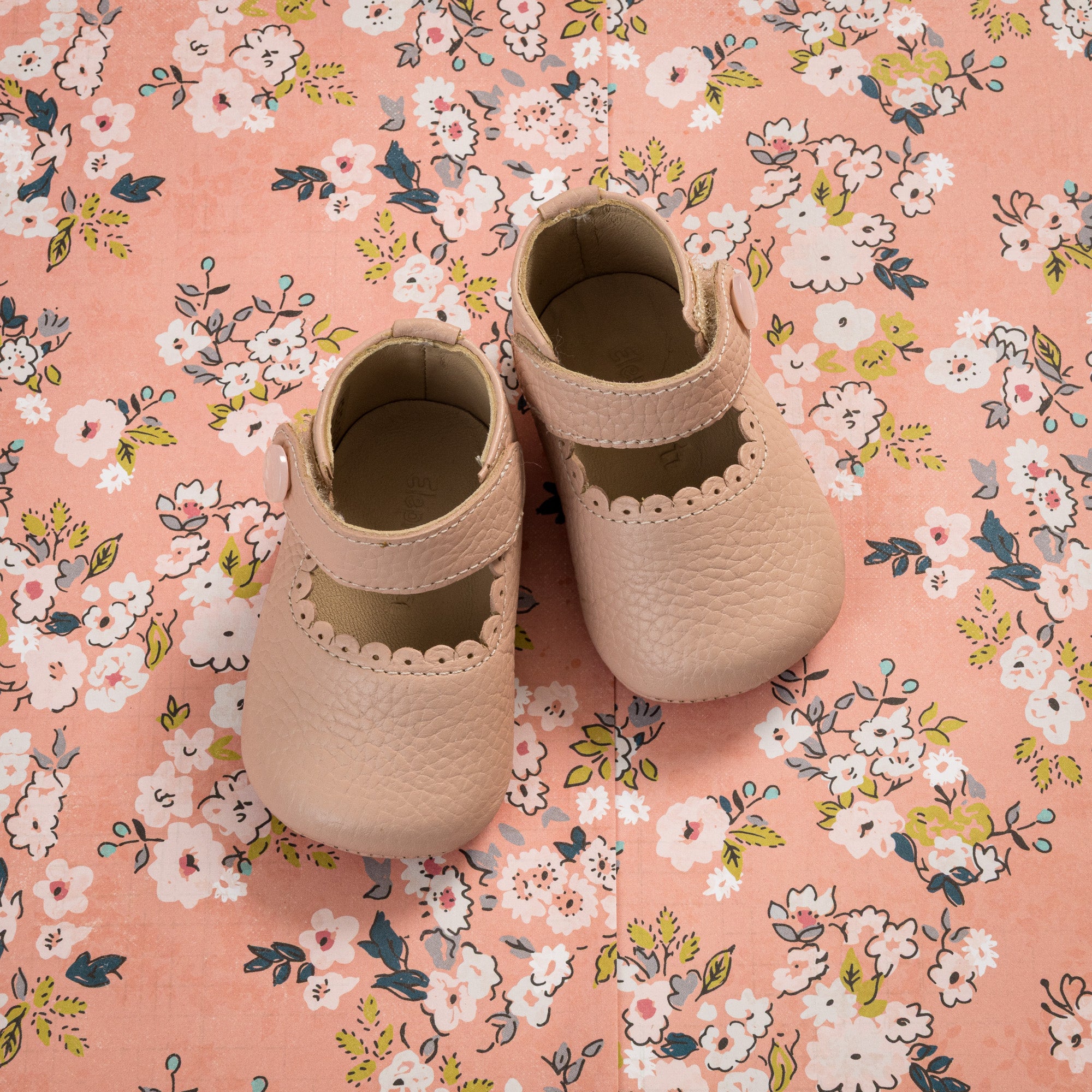 Soft pink baby Mary Jane shoes with scalloped edges on floral fabric background.