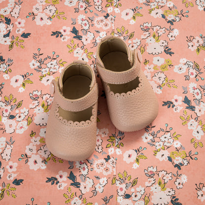 Soft pink baby Mary Jane shoes with scalloped edges on floral fabric background.
