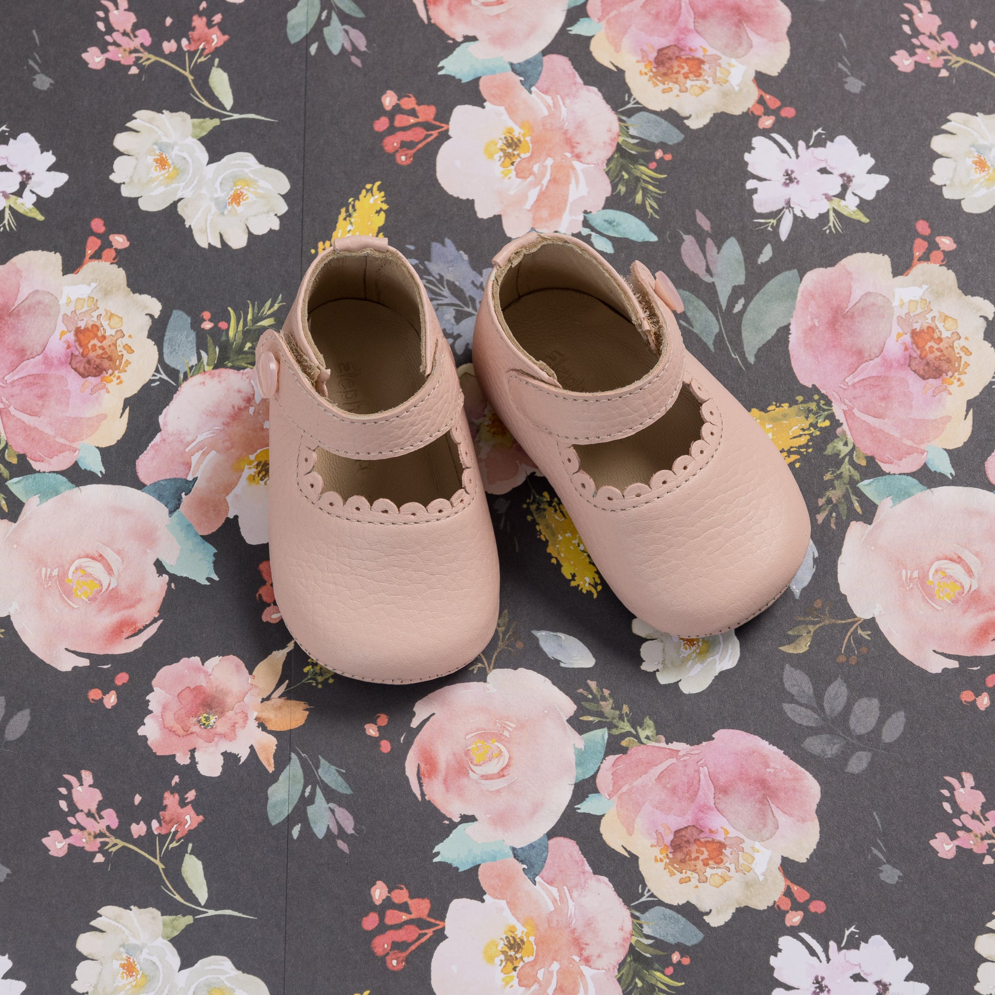 Soft pink baby Mary Jane shoes with scalloped edges on floral background.