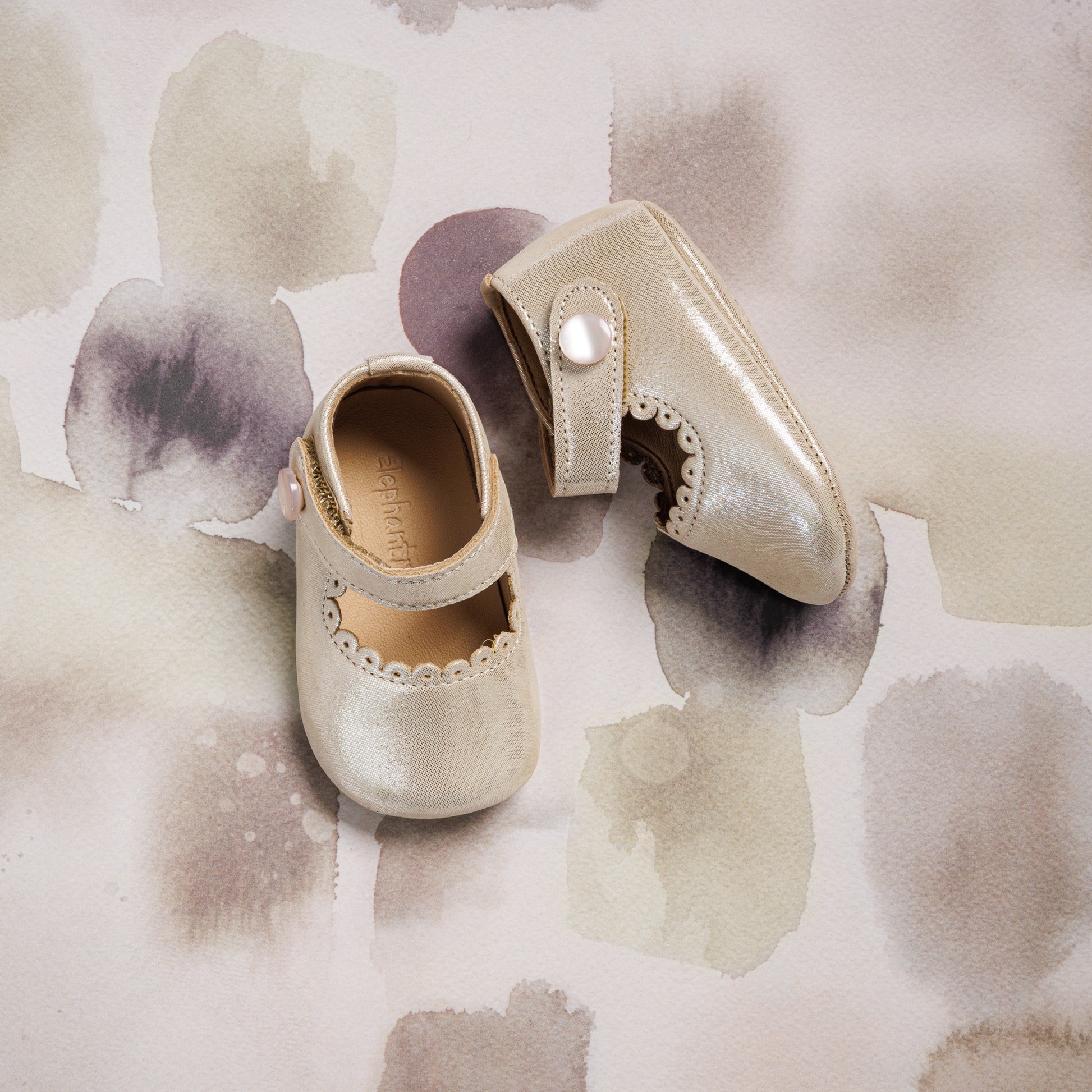 Cream-colored baby Mary Jane shoes with scalloped edges and pearl button straps on a pastel watercolor background.