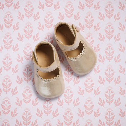 Beige toddler Mary Jane shoes with scalloped edge and velcro strap on patterned pink background.