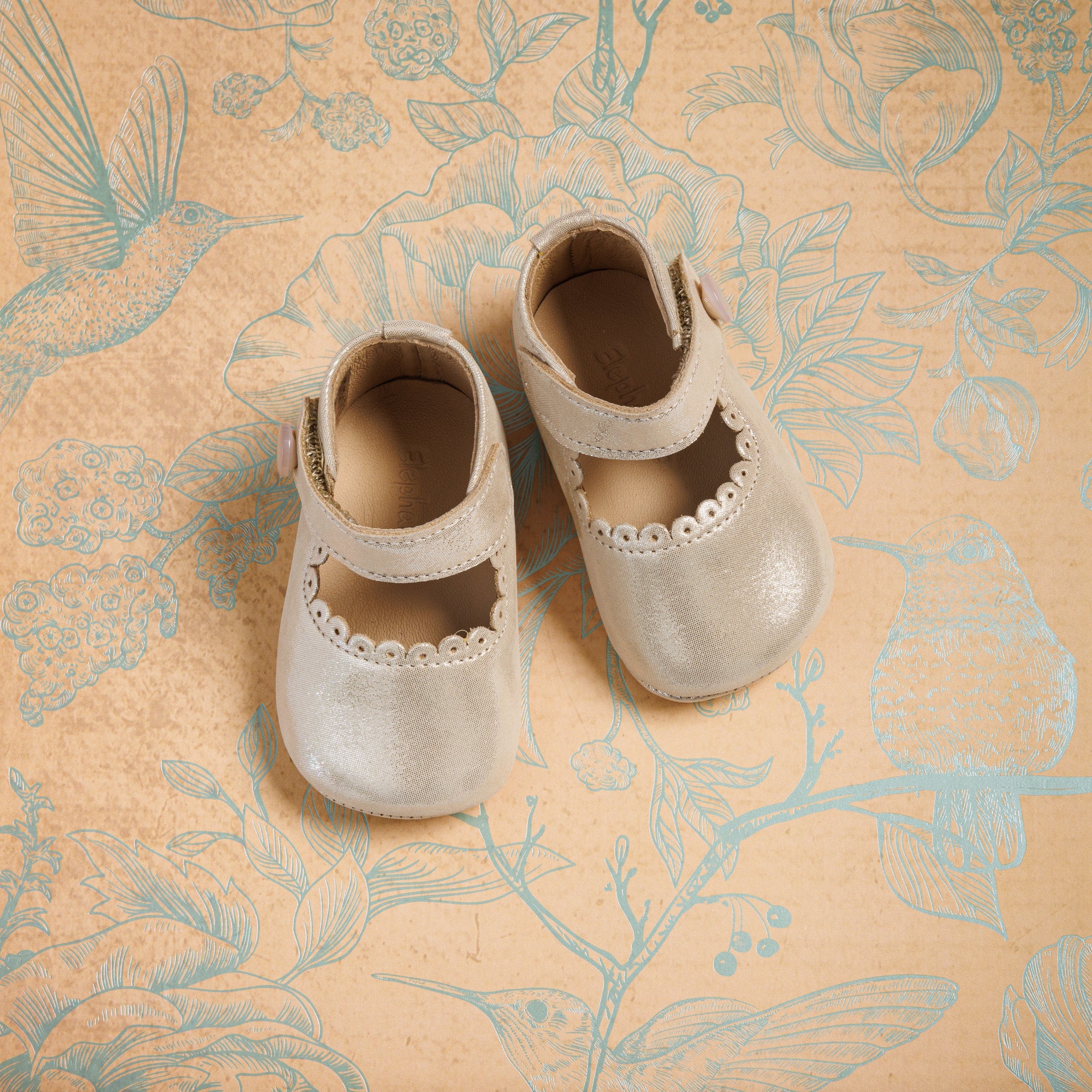 Soft beige toddler Mary Jane shoes with scalloped edges and Velcro straps on patterned fabric background.