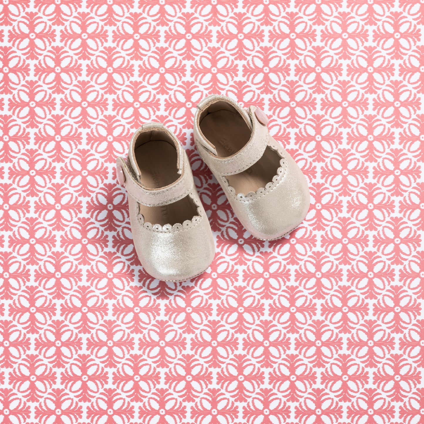 White baby Mary Jane shoes with scalloped edges and a soft pink strap on pink patterned background.