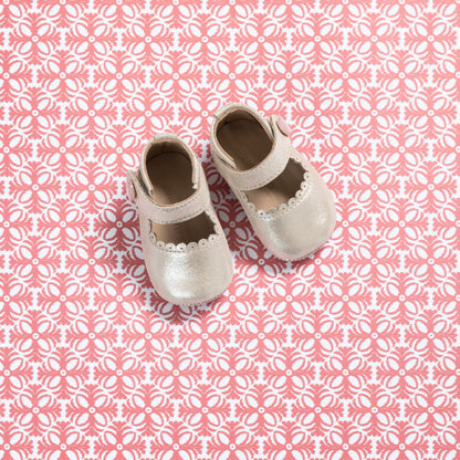 White baby Mary Jane shoes with scalloped edges and a soft pink strap on pink patterned background.
