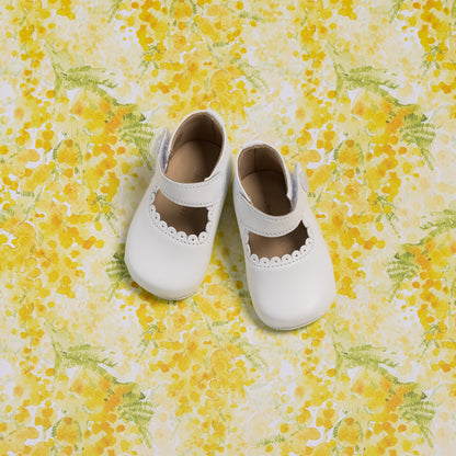 White baby Mary Jane shoes with scalloped edge and strap on yellow floral background.