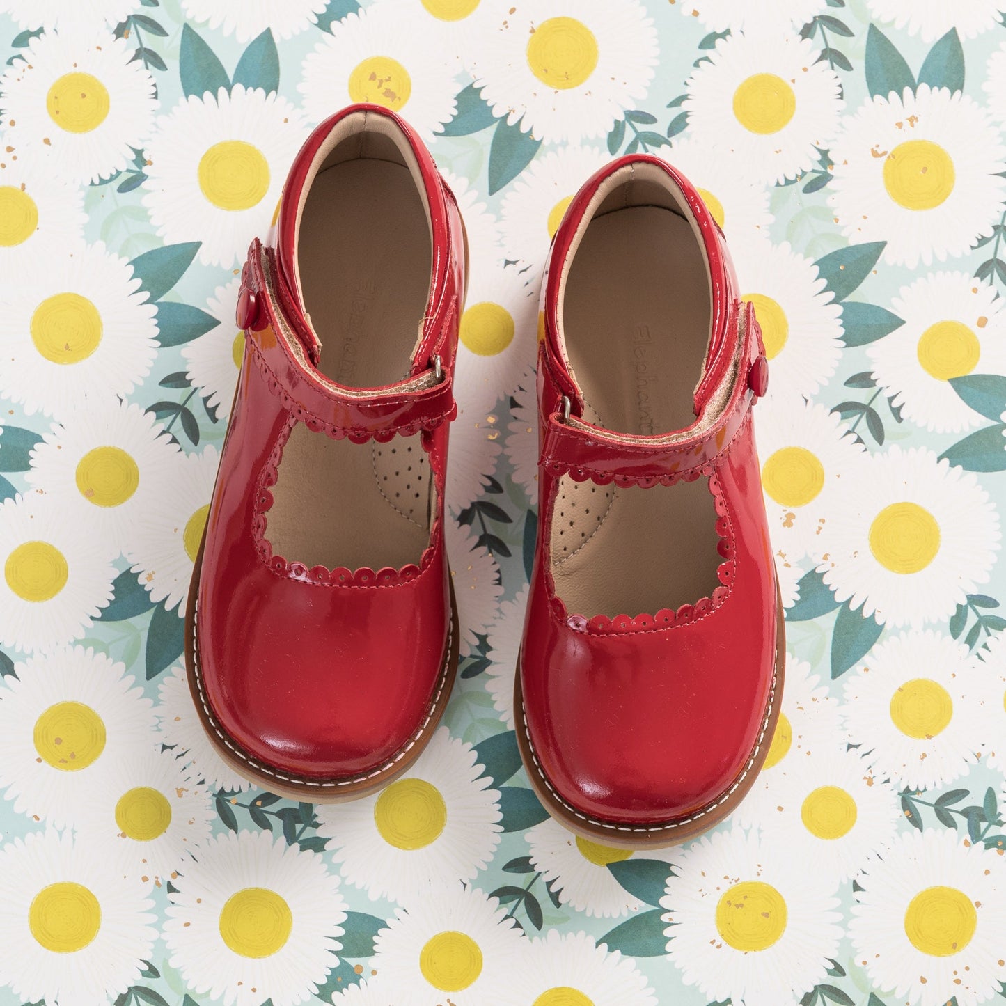 Shiny red patent leather Mary Jane shoes with scalloped edges and velcro straps on floral background.