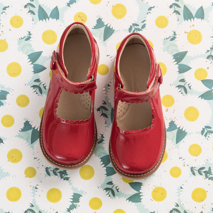 Shiny red patent leather Mary Jane shoes with scalloped edges and velcro straps on floral background.