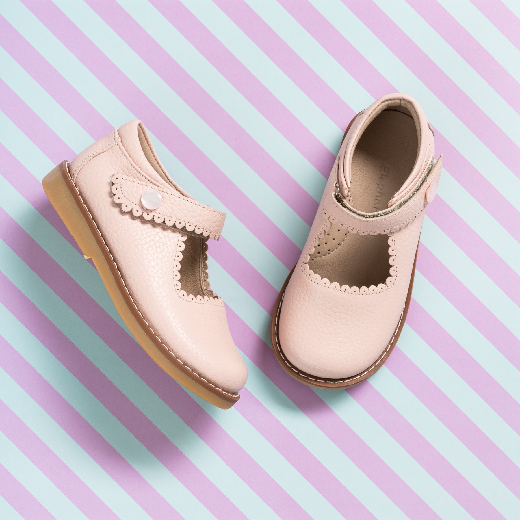 Light pink leather girls’ Mary Jane shoes with scalloped strap and tan soles on striped pastel background.