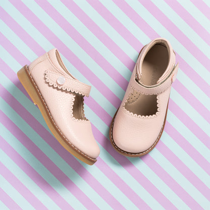 Light pink leather girls’ Mary Jane shoes with scalloped strap and tan soles on striped pastel background.