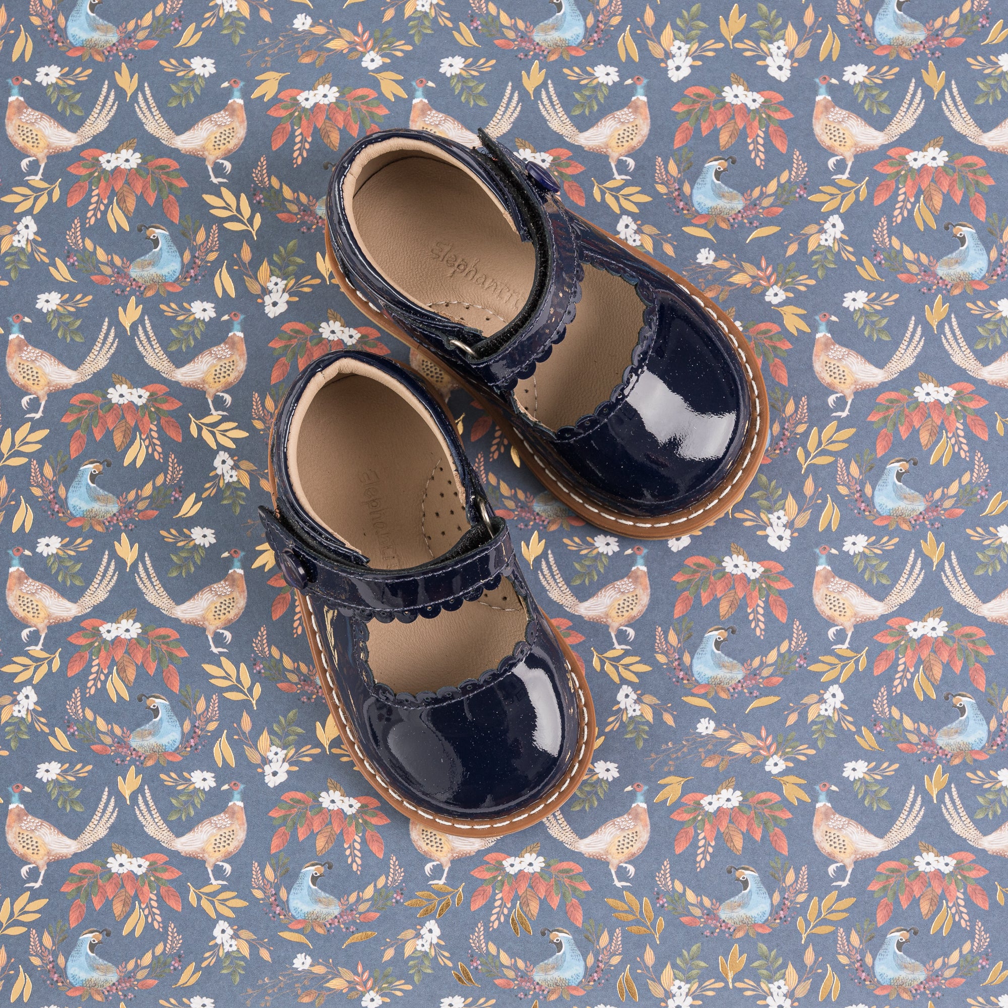 Navy blue patent leather toddler Mary Jane shoes with scalloped edge and strap closure on floral background.