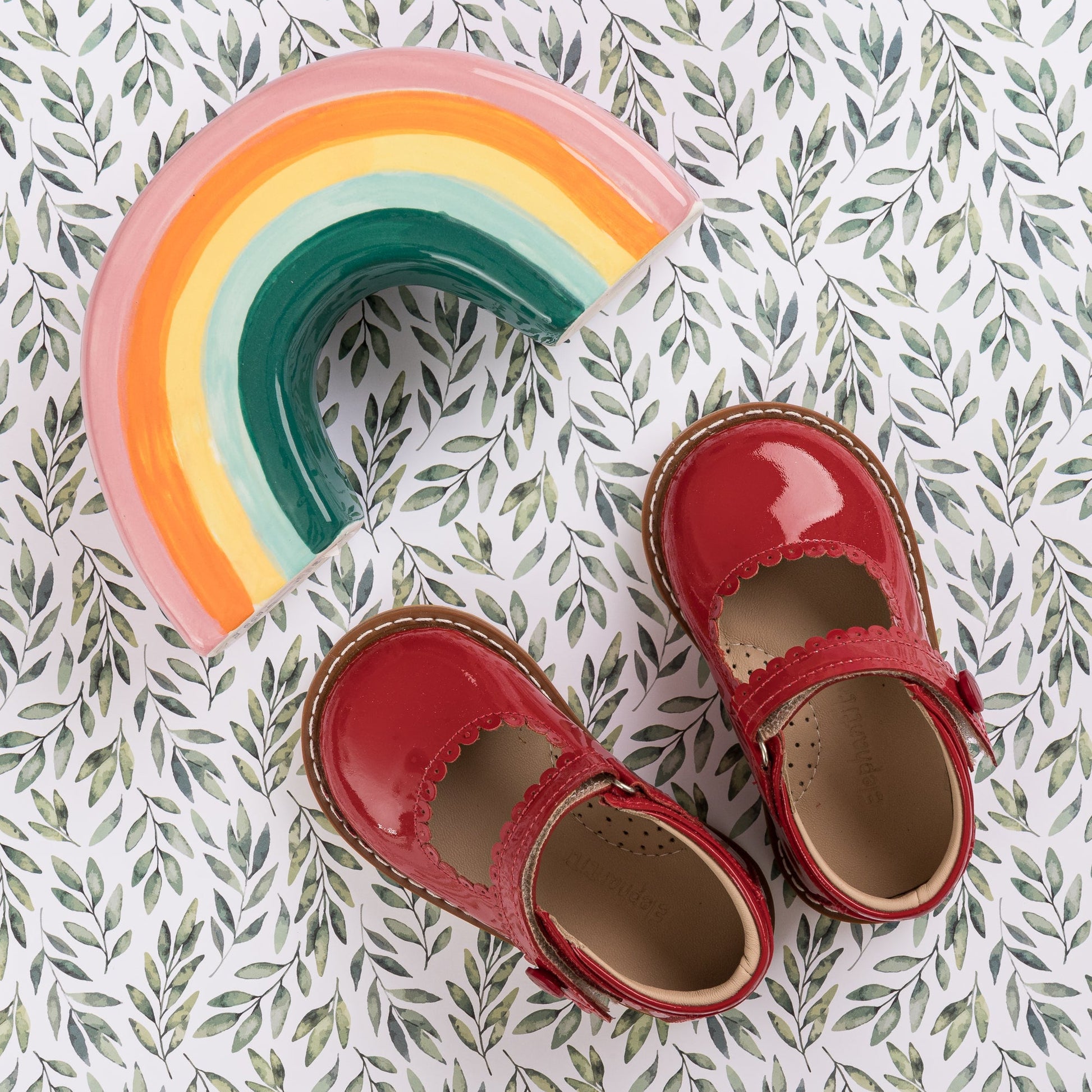 Glossy red patent Mary Jane toddler shoes with scalloped edges and adjustable straps on leafy background.