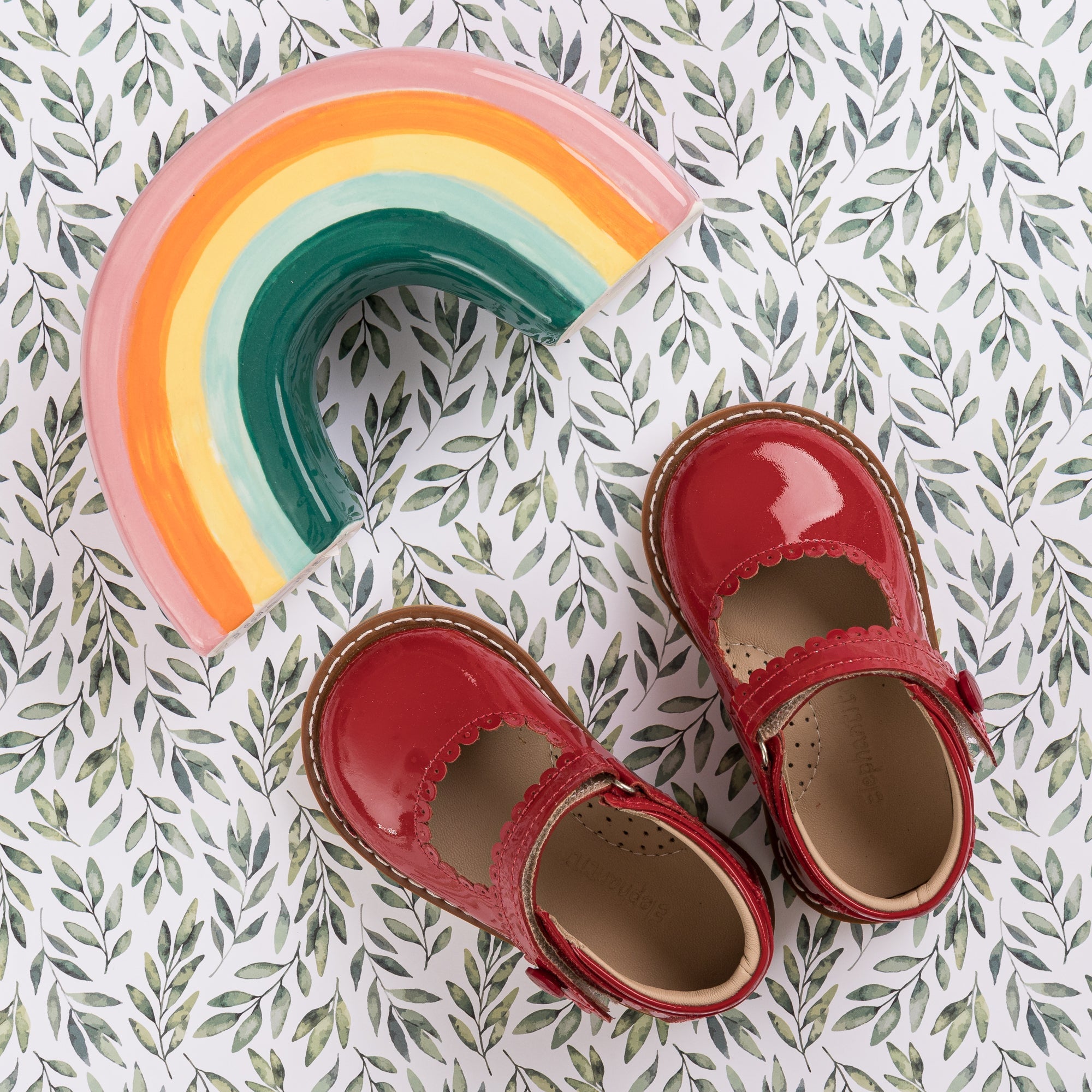 Glossy red patent Mary Jane toddler shoes with scalloped edges and adjustable straps on leafy background.