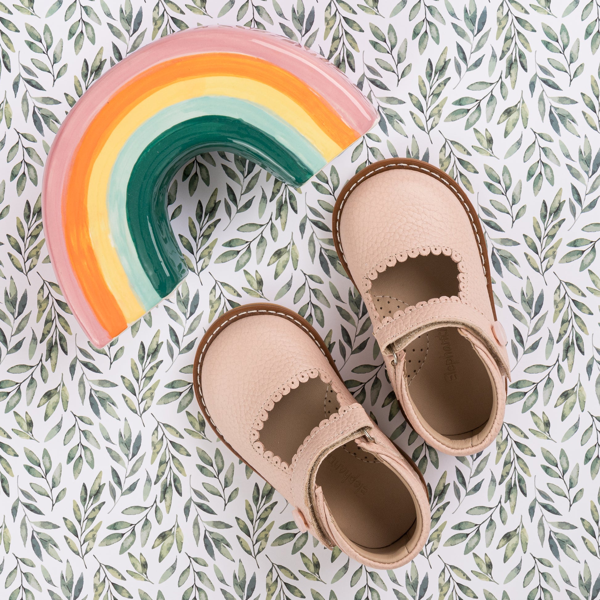 Pink toddler Mary Jane shoes with scalloped edges on leafy fabric background.