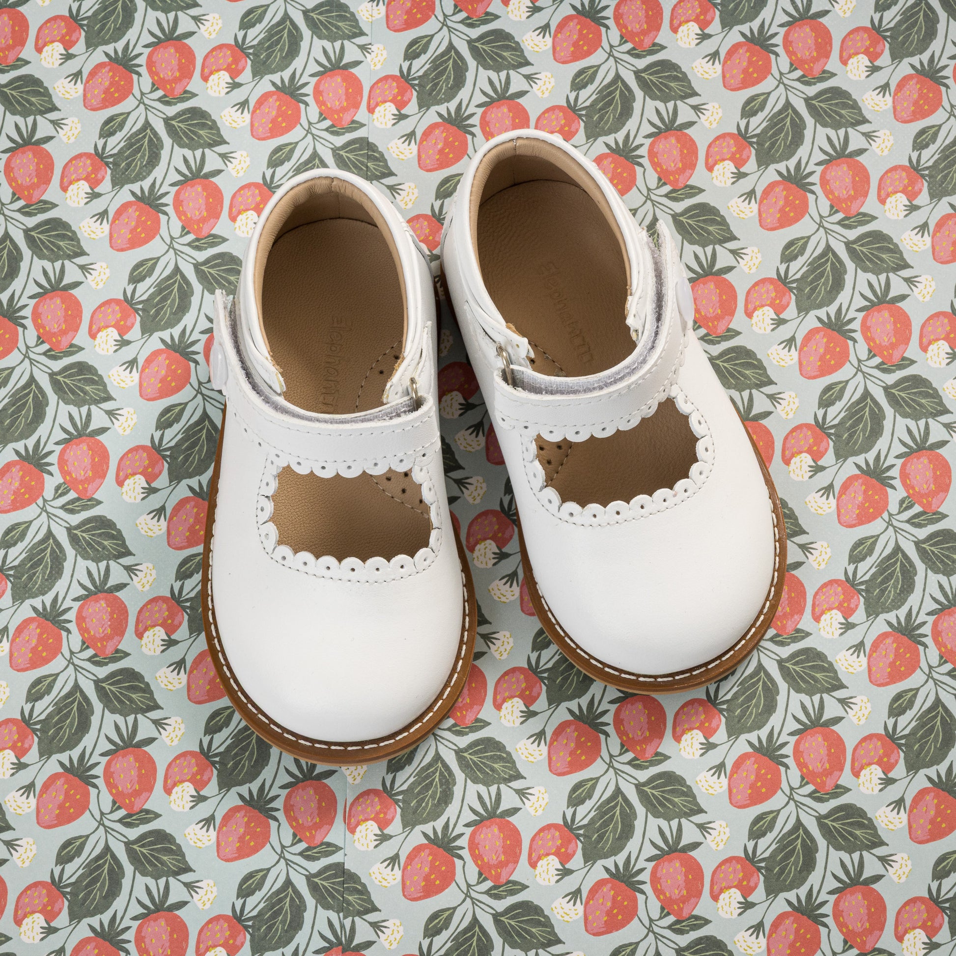 White toddler Mary Jane shoes with scalloped edges and brown soles on floral fabric background.
