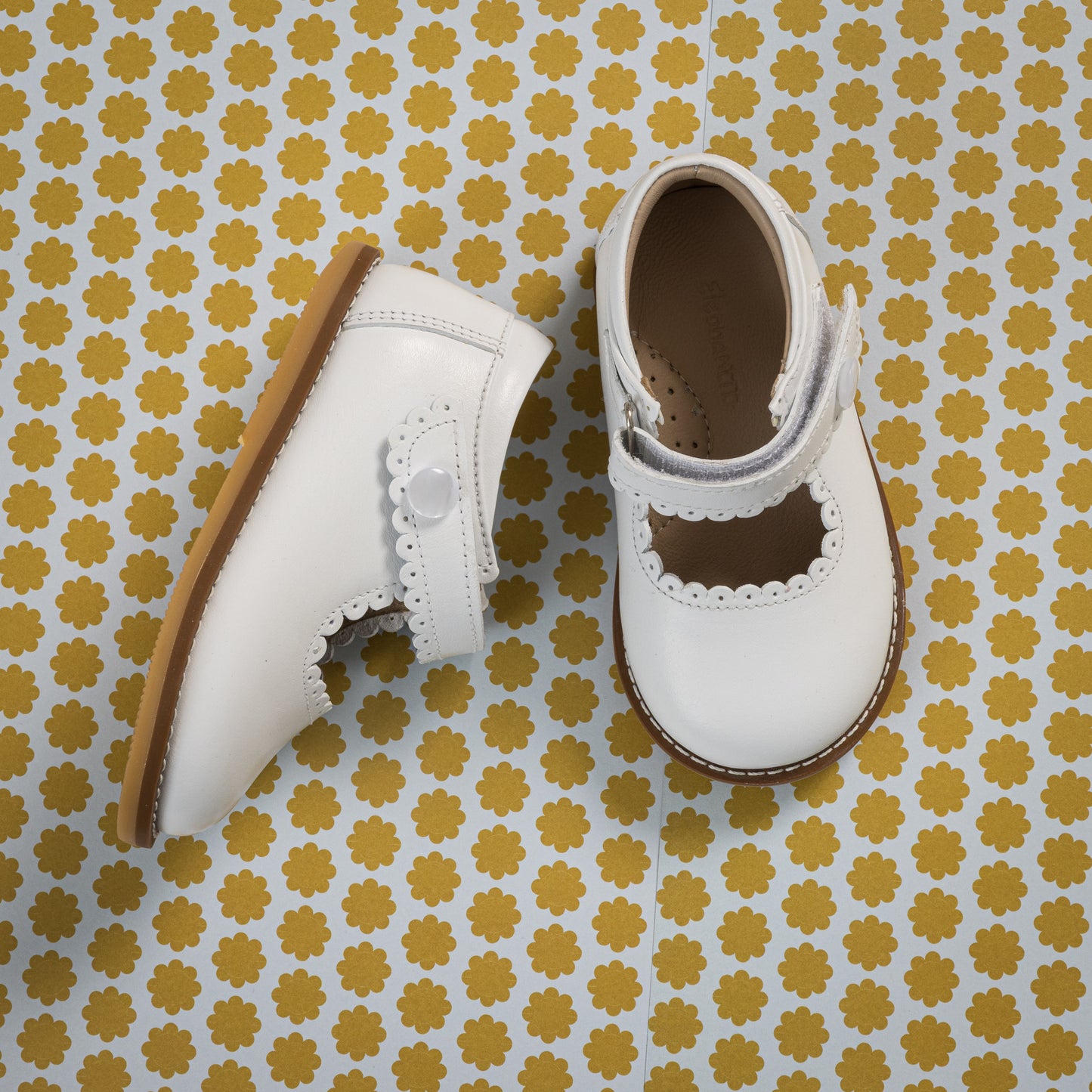 White toddler Mary Jane shoes with scalloped edges and Velcro strap on patterned yellow background.