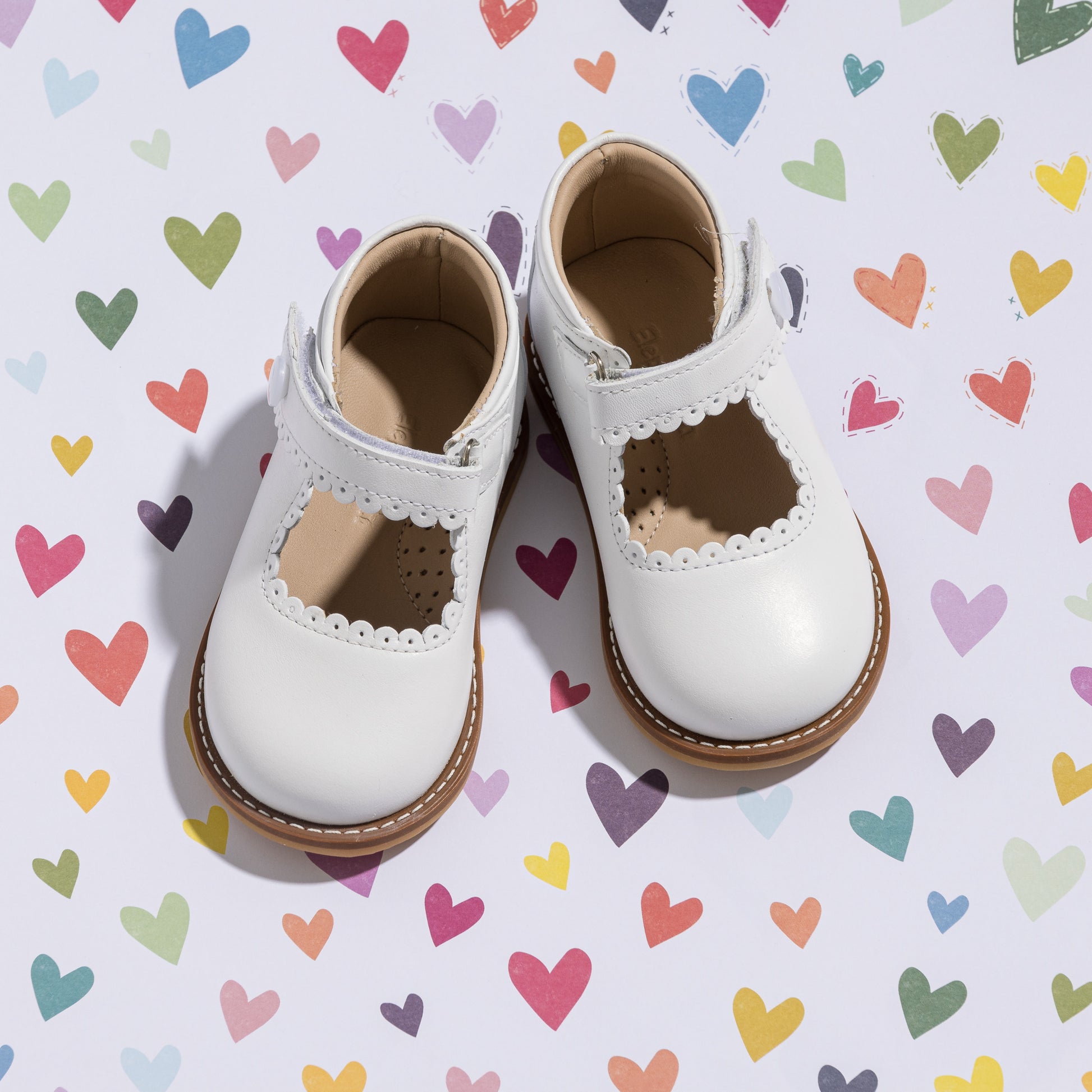 White toddler Mary Jane shoes with scalloped edges and brown soles on colorful heart background.