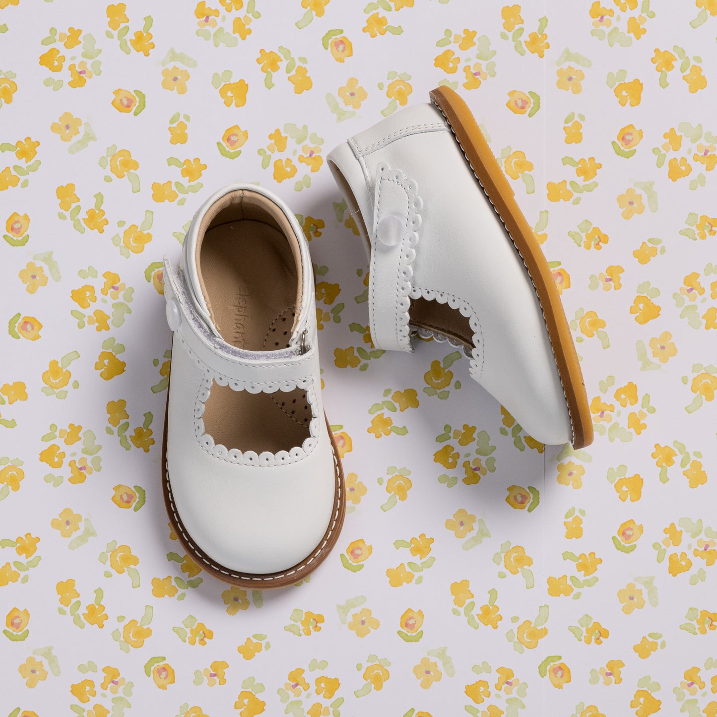 White toddler Mary Jane shoes with scalloped edges and a brown sole on floral background.