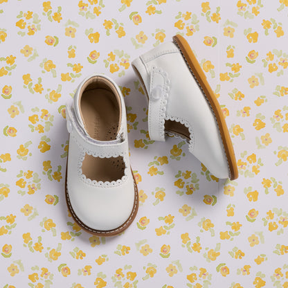 White toddler Mary Jane shoes with scalloped edges and a brown sole on floral background.