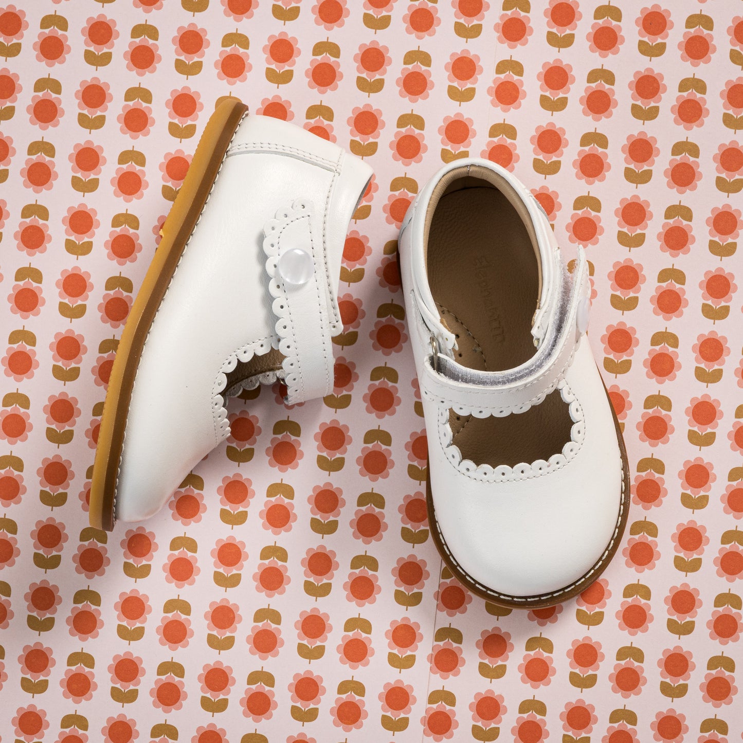 White toddler Mary Jane shoes with scalloped edges and tan soles on patterned background.
