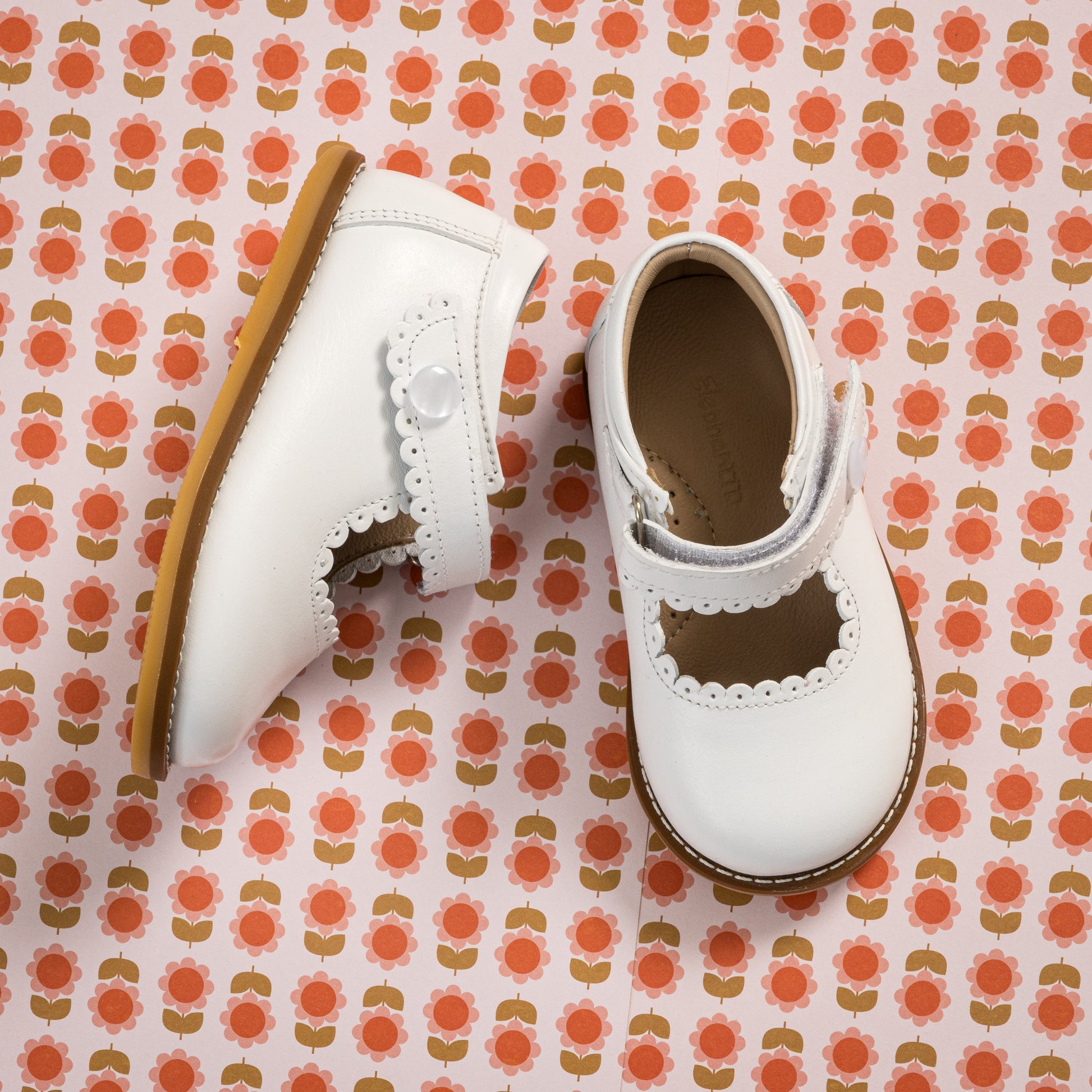 White toddler Mary Jane shoes with scalloped edges and tan soles on patterned background.