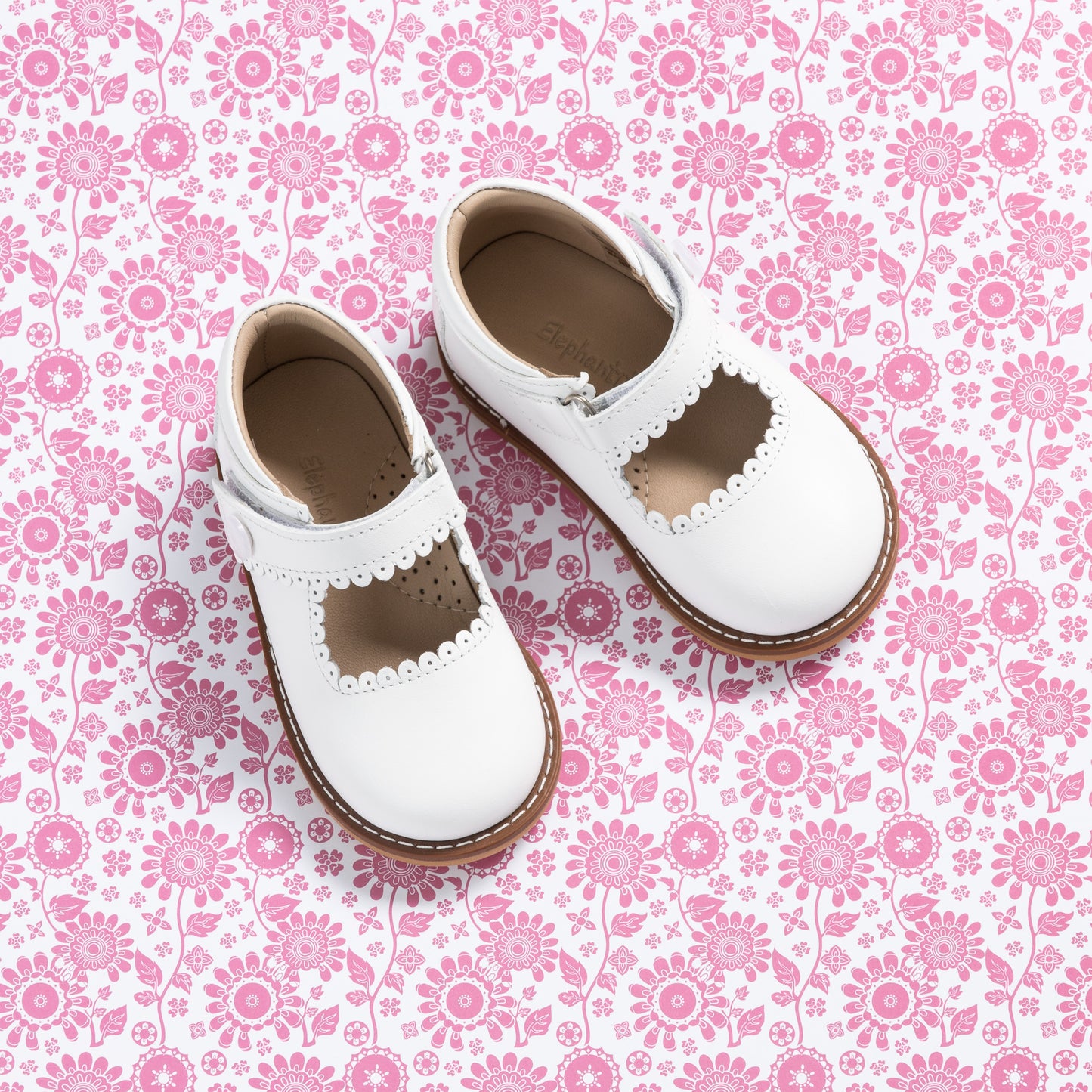 White leather girls' Mary Jane shoes with scalloped edges and brown rubber soles on pink floral background.