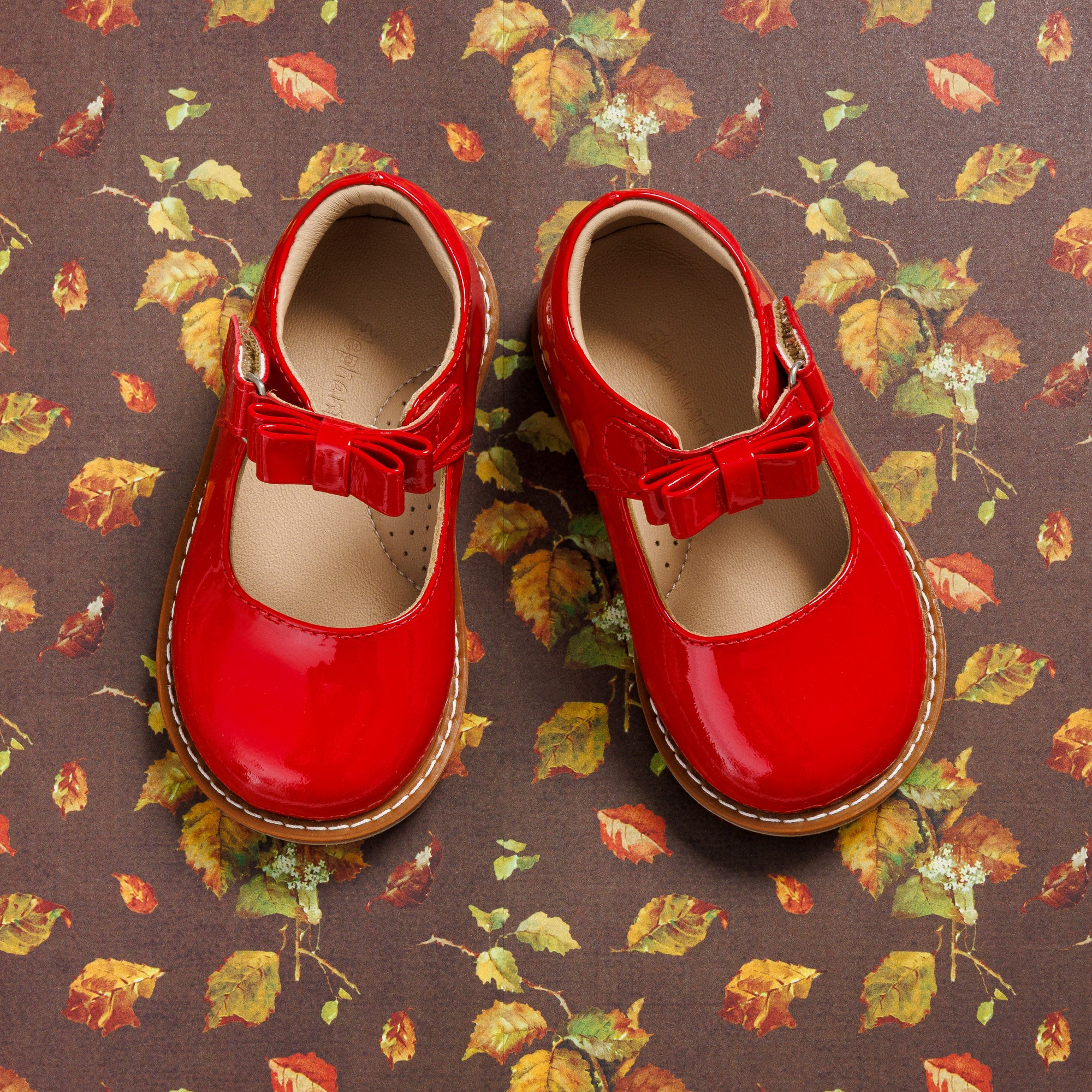 Glossy red toddler Mary Jane shoes with bow accents and tan stitching on a floral background.