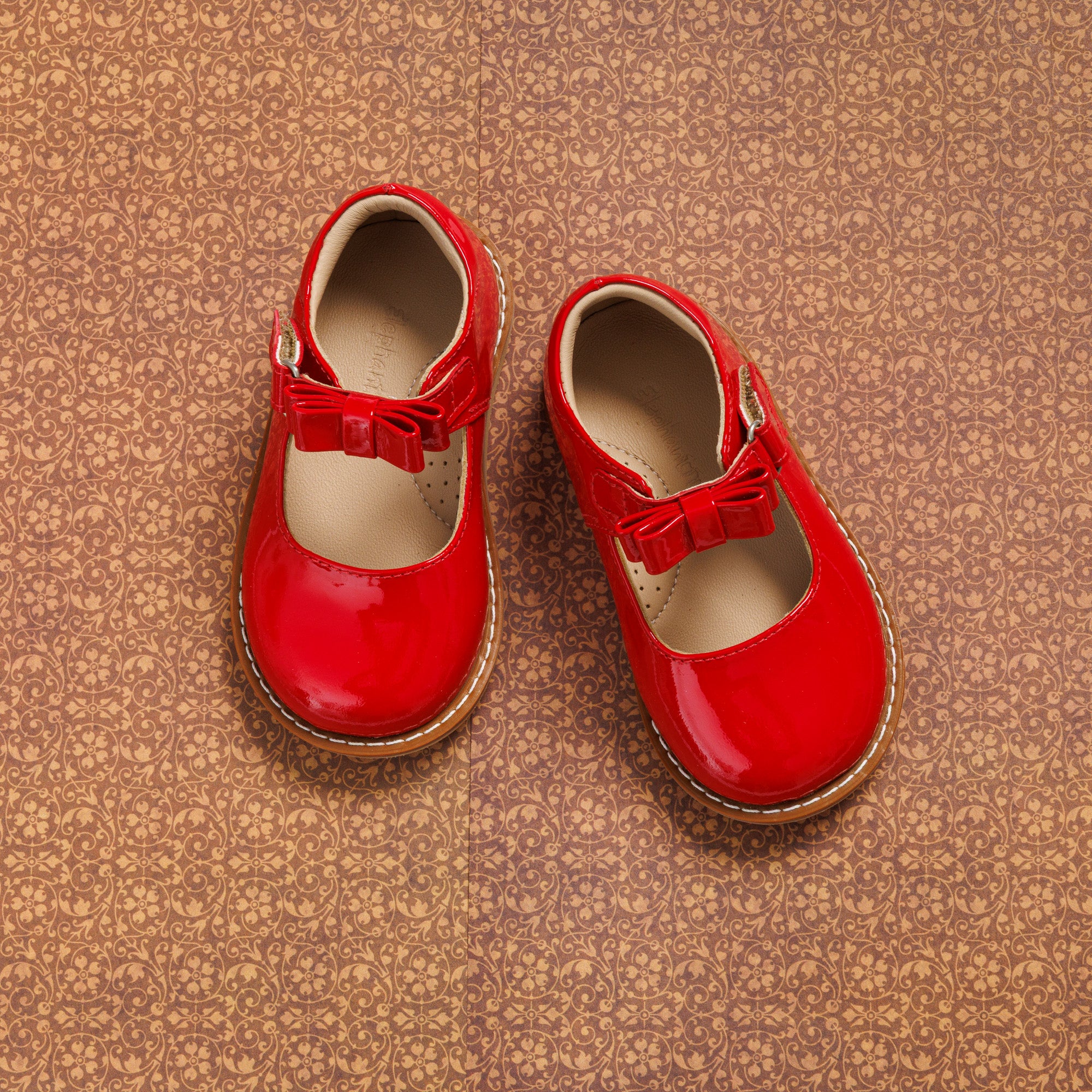 Glossy red toddler Mary Jane shoes with a bow detail and brown soles on a textured background.