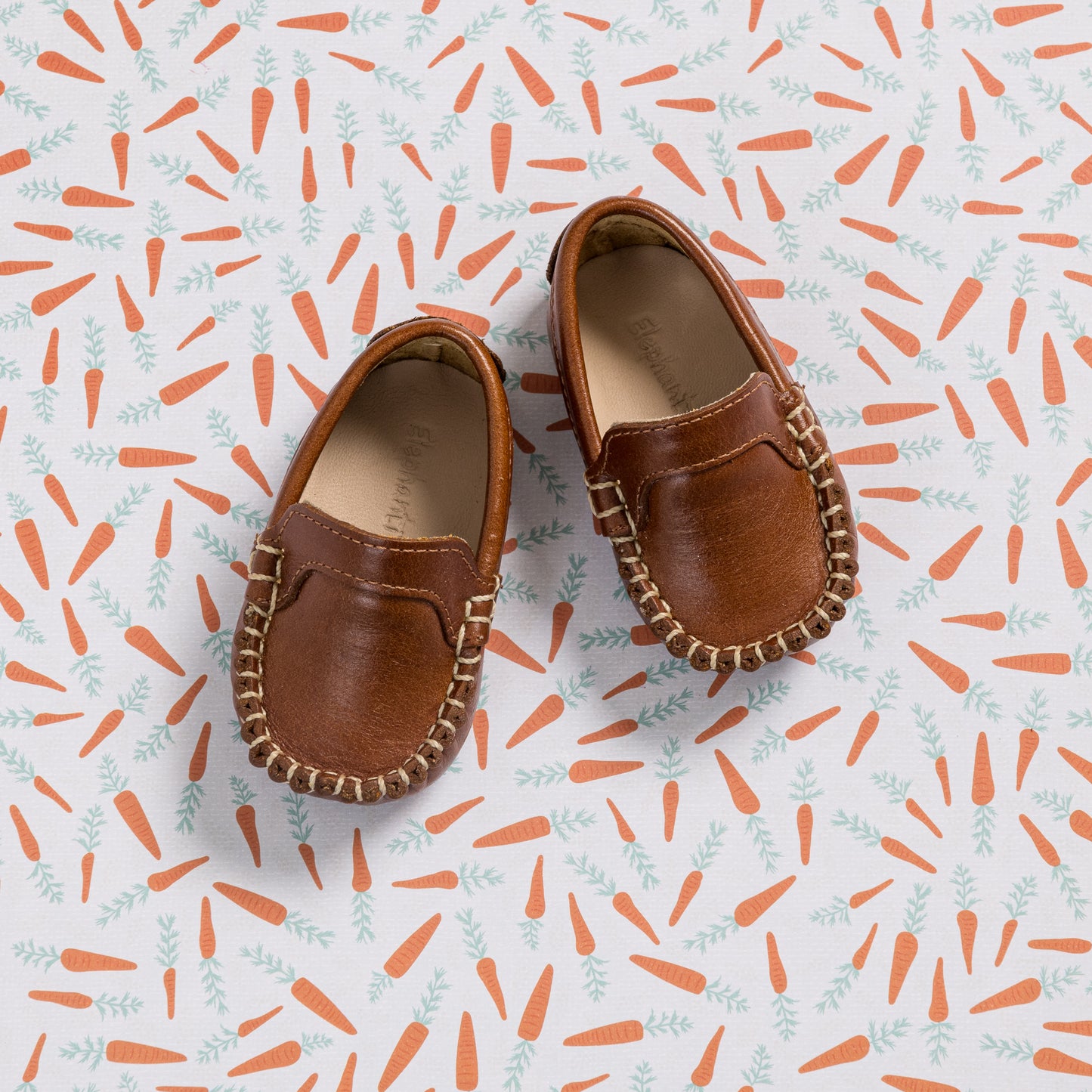 Natural brown leather baby moccasins with stitched detailing on a patterned background.
