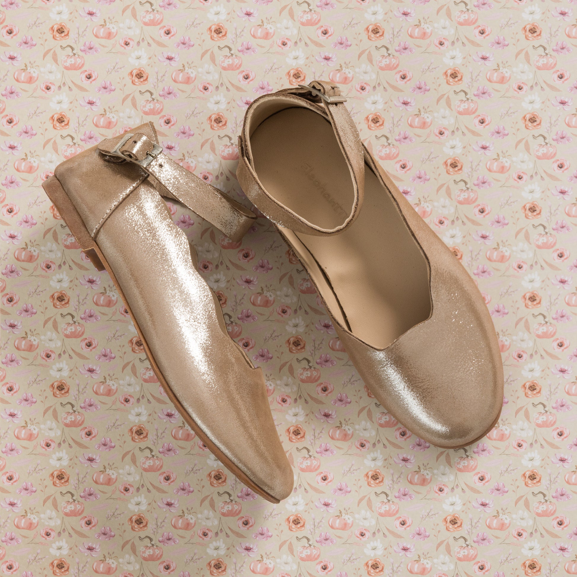 Blush pink toddler ballerina flats with ankle strap and subtle sheen on floral background.