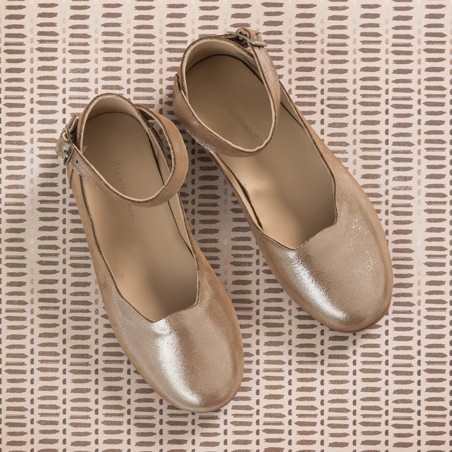 Shiny blush pink toddler ballerina flats with ankle straps on textured beige background.