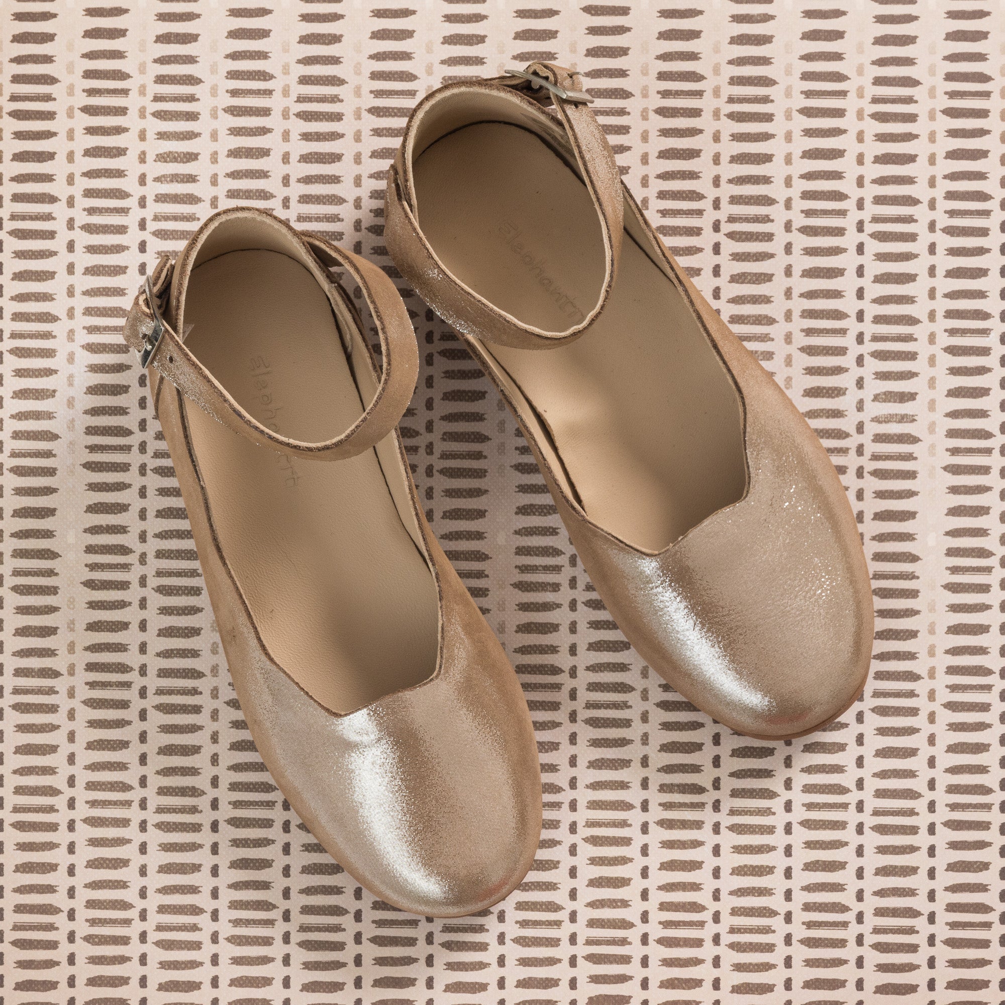 Shiny blush pink toddler ballerina flats with ankle straps on textured beige background.