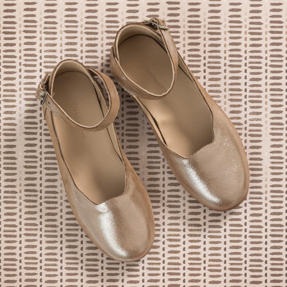 Shiny blush pink toddler ballerina flats with ankle straps on textured beige background.
