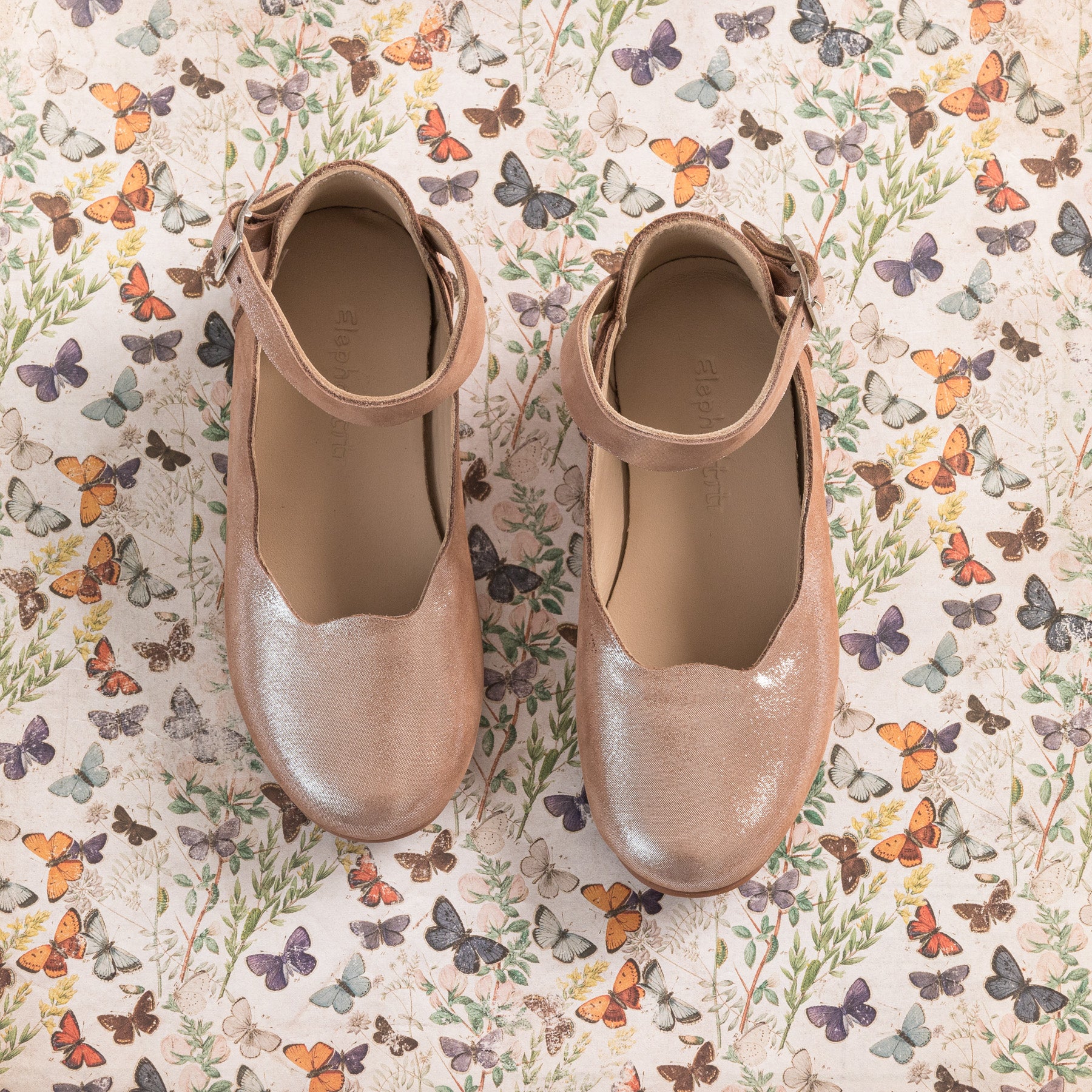 Blush pink toddler ballerina flats with ankle straps on butterfly print background.