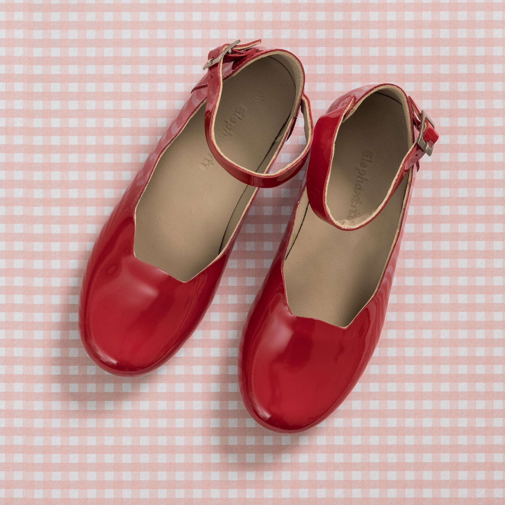 Shiny red patent leather girls' ballerina flats with ankle straps on pink gingham background.
