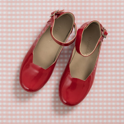 Shiny red patent leather girls' ballerina flats with ankle straps on pink gingham background.
