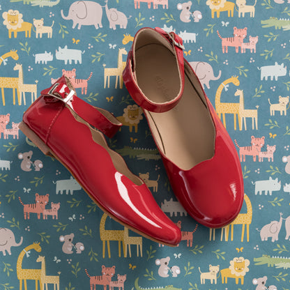 Glossy red patent leather ballerina flats with ankle strap and buckle on colorful animal print background.