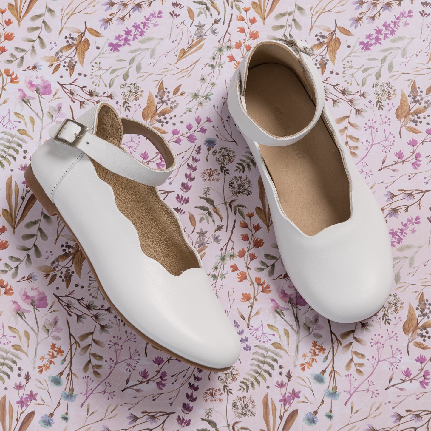White leather girls' ballerina flats with scalloped edges and ankle buckle straps on floral background.