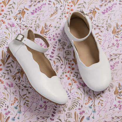 White leather girls' ballerina flats with scalloped edges and ankle buckle straps on floral background.